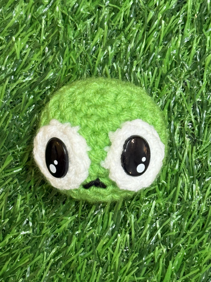 A picture of Steve the pea-shaped Chunkle Puff with large black eyes and a small frown, made from green and white yarn, resting on green grass.