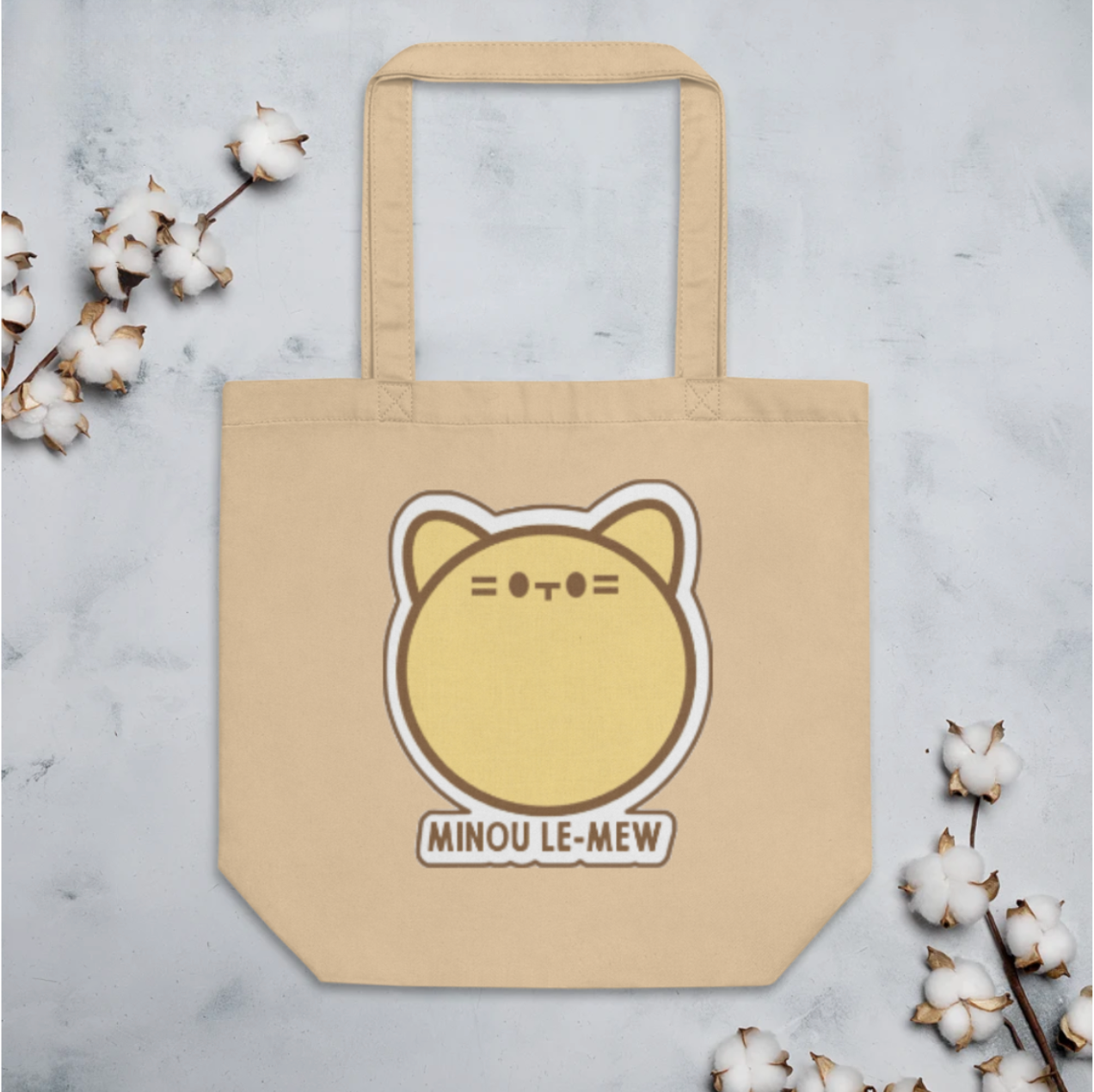 Beige tote bag with a cat illustration and 'MINOU LE-MEW' text on a light gray background with cotton branches.