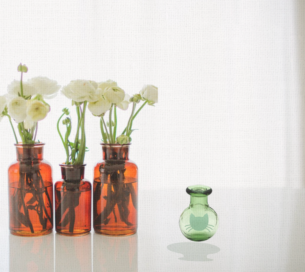 A green glass Whisker Vase with foil cat silhouette displayed beside amber glass vials filled with flowers, styled as a boutique keepsake.